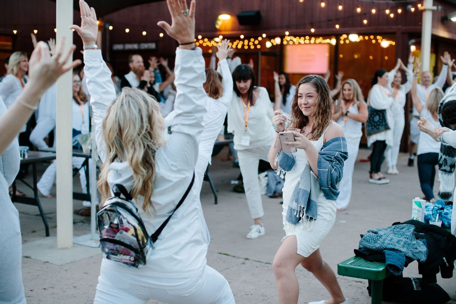 Crowd of participants in white clothing gathering outdoors under string lights at dusk.