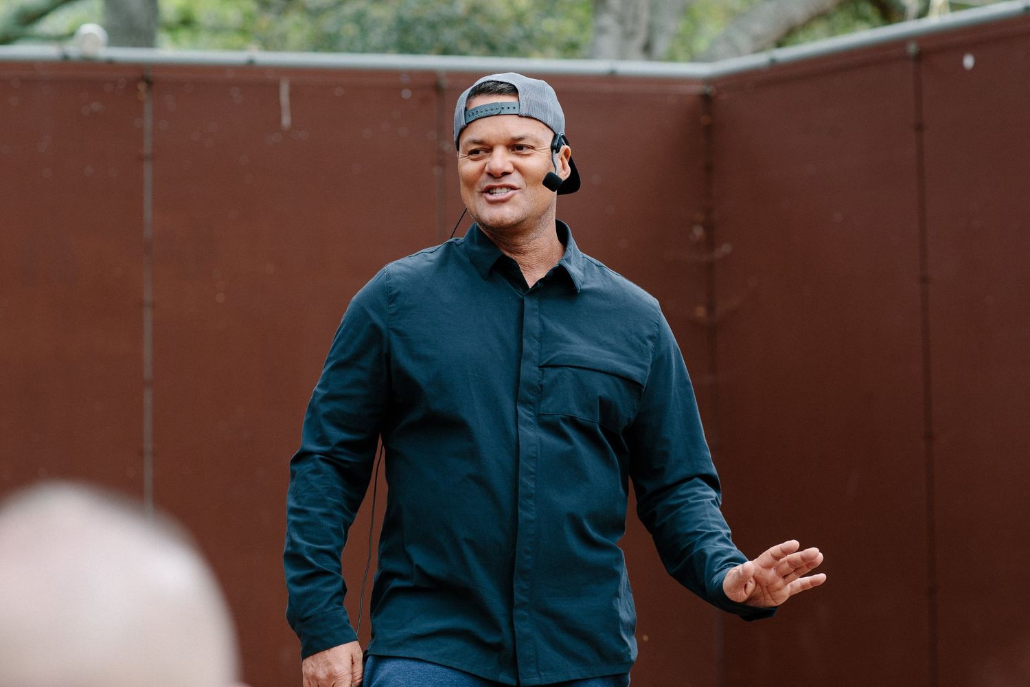 Speaker engaging with audience outdoors against a rustic brown wall background.