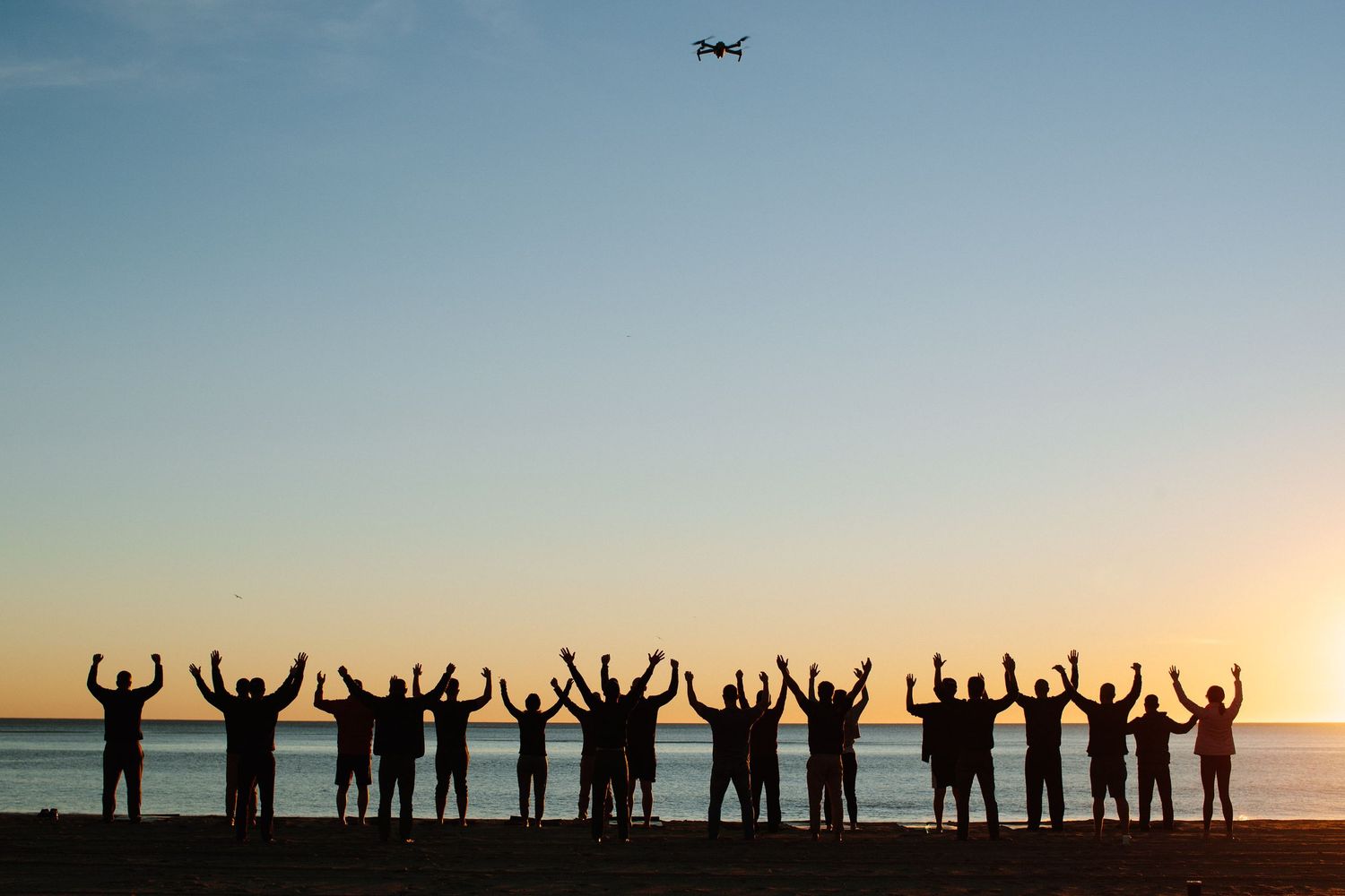 Silhouettes of people raising arms against sunset sky with drone flying overhead.