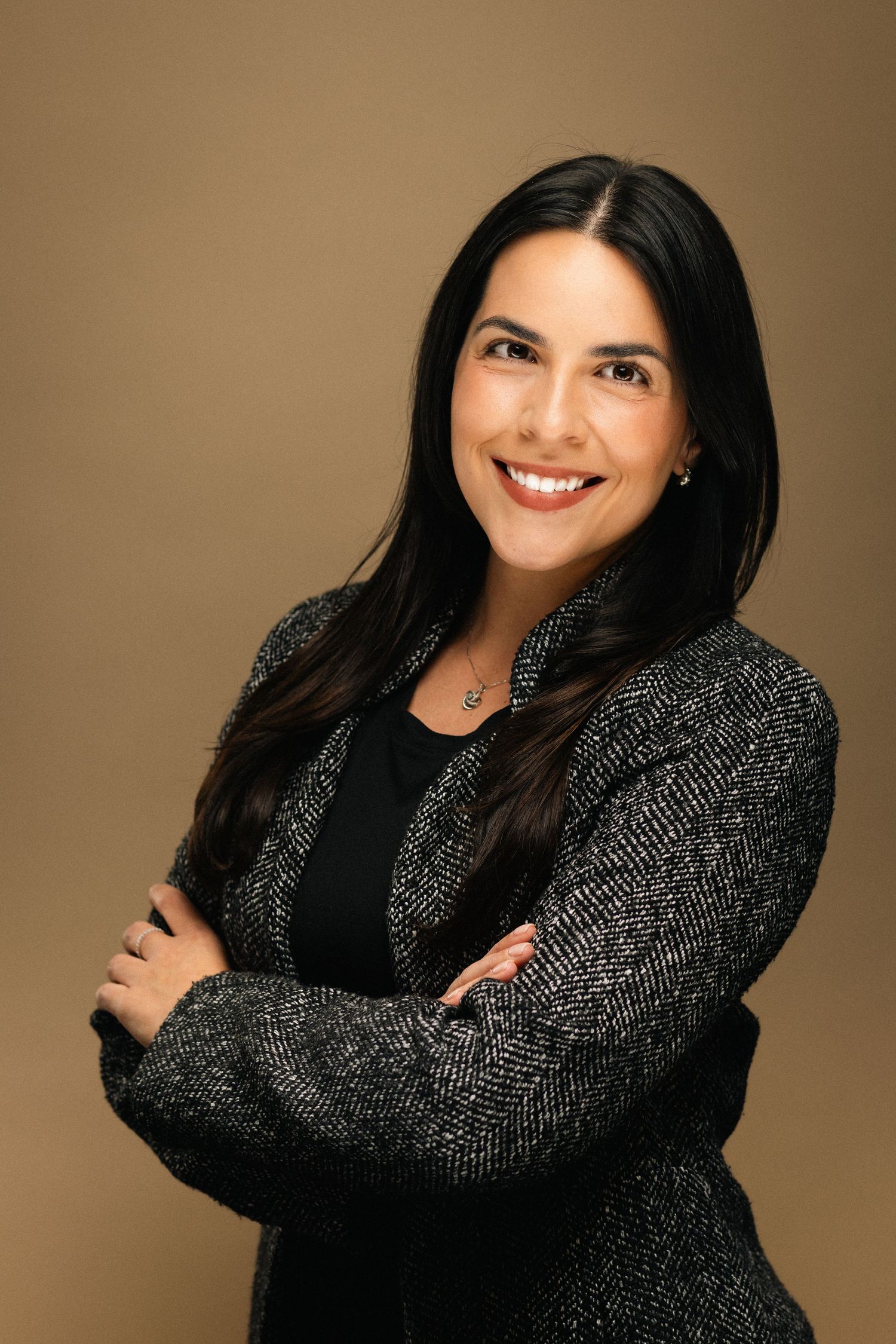 Professional woman with dark hair smiling confidently, arms crossed, wearing a gray blazer against a brown background.