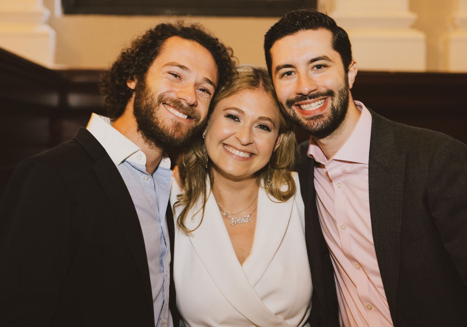 Three friends share a warm embrace while dressed in formal attire at an indoor celebration.