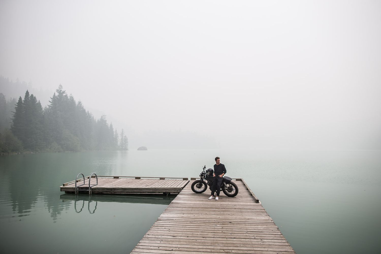 A silhouetted motorcyclist stands on a misty wooden dock overlooking a tranquil foggy lake surrounded by evergreen trees.