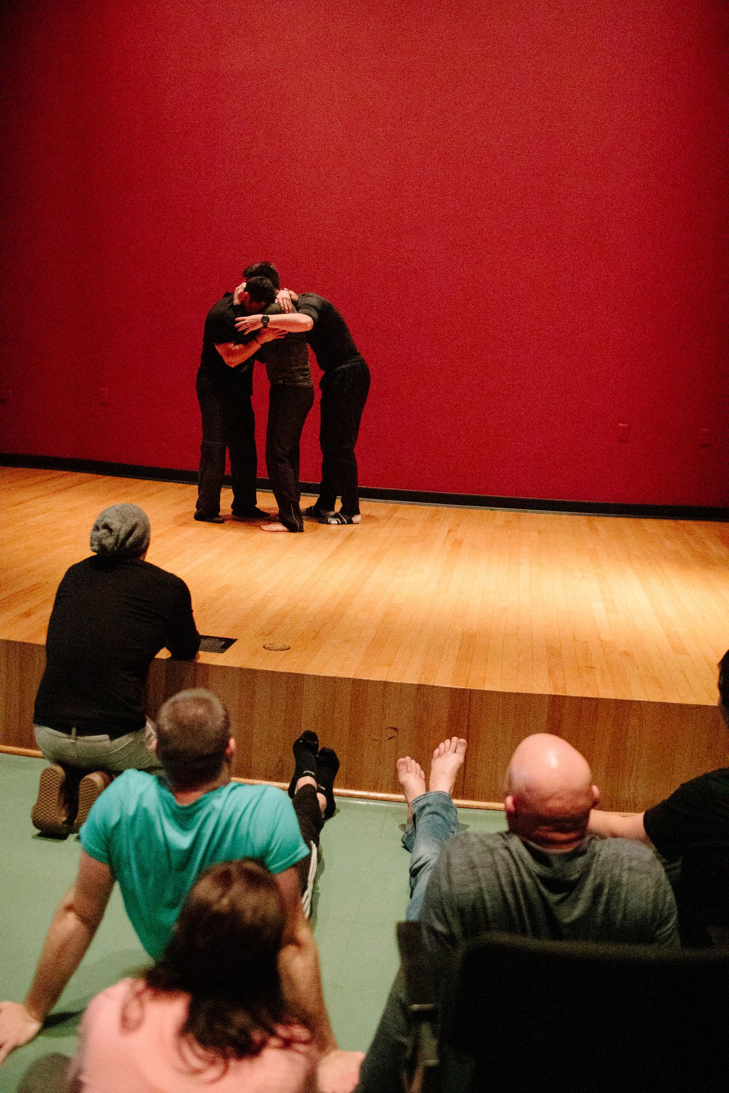 Several people seated in a theater space with red walls and wooden flooring.