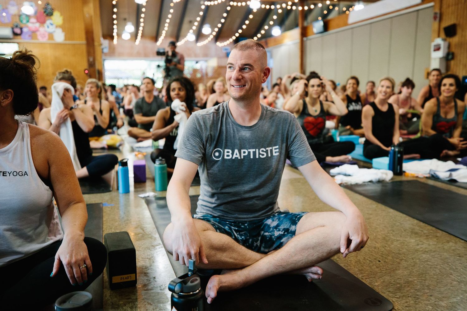 Large yoga class with students sitting in meditation pose on mats while an instructor demonstrates at the front.