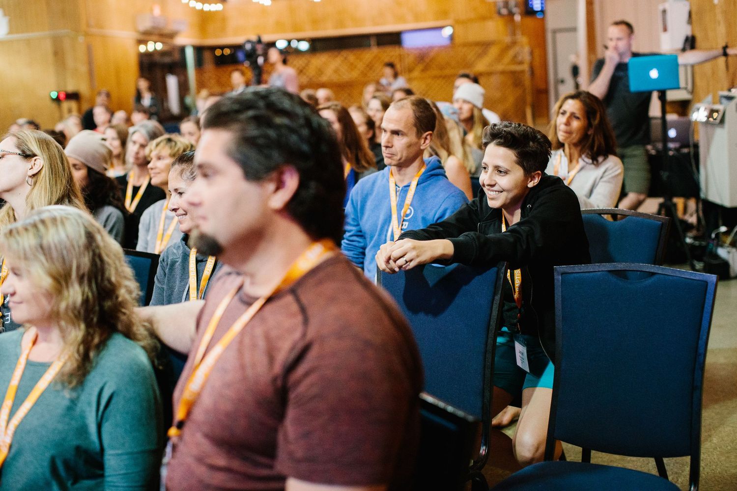 Row of attendees sitting in blue chairs during an indoor conference or event.