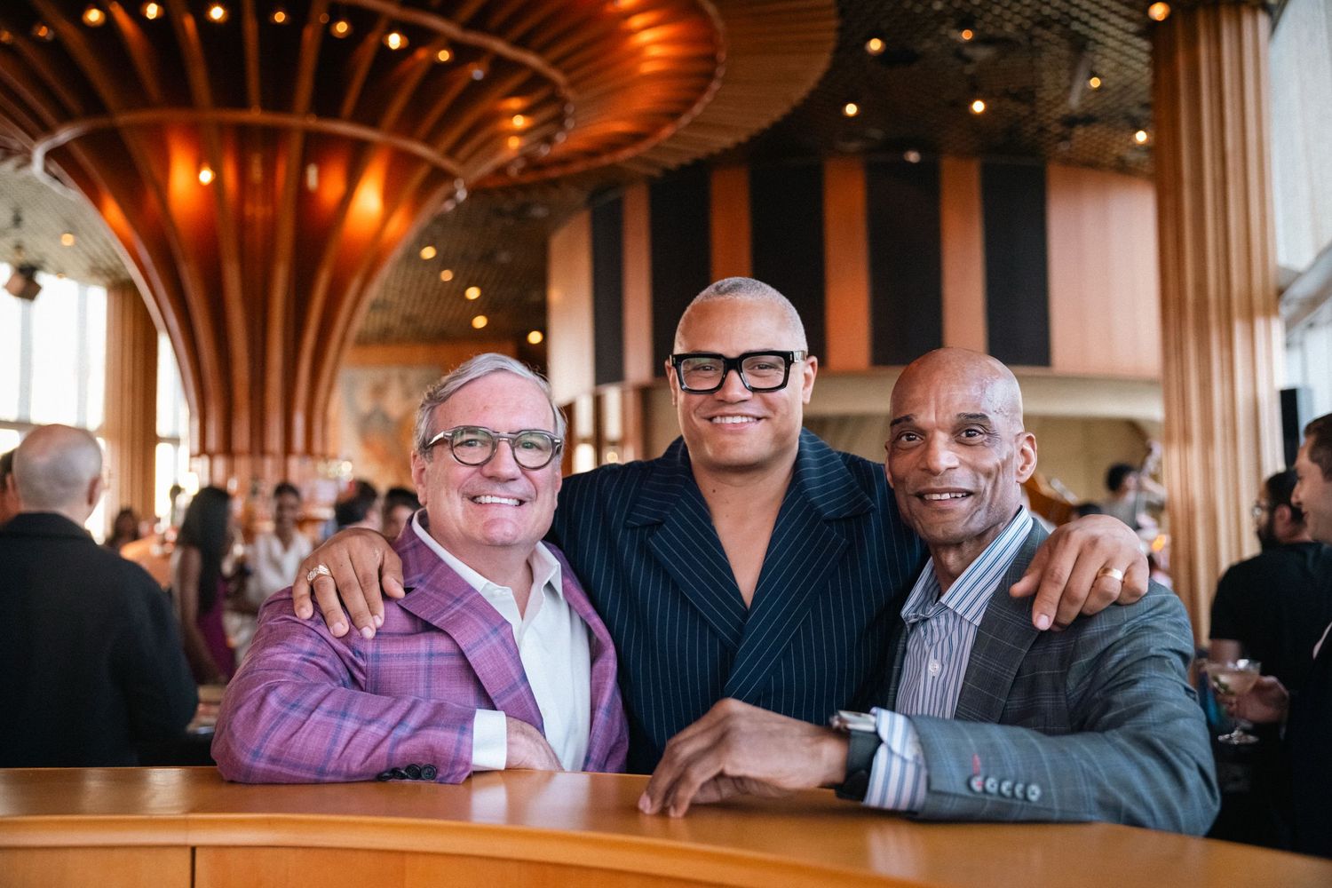 Three smiling men pose together at a restaurant bar, dressed in stylish blazers in a warmly lit venue.