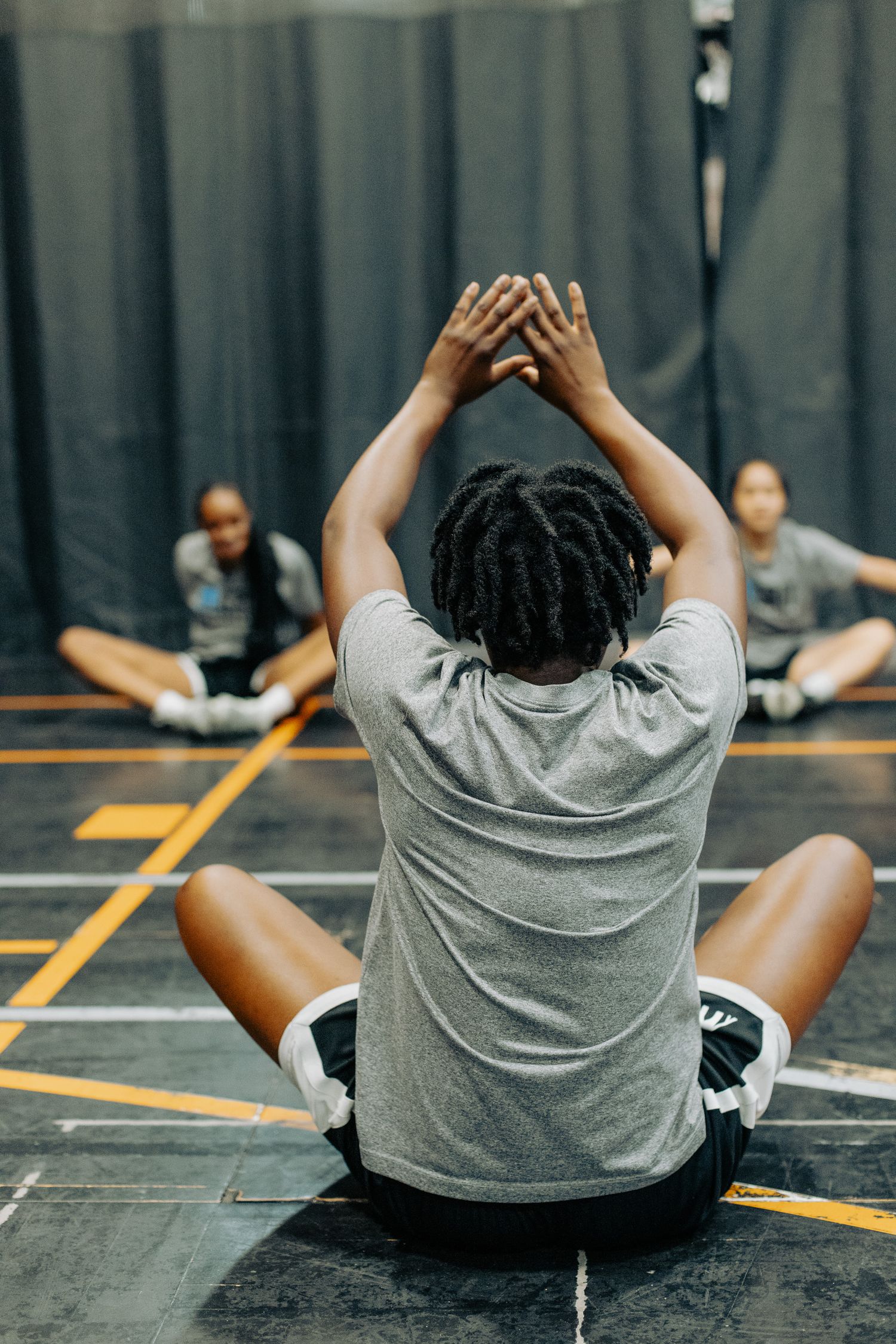 Player stretches on indoor basketball court with arms raised overhead.