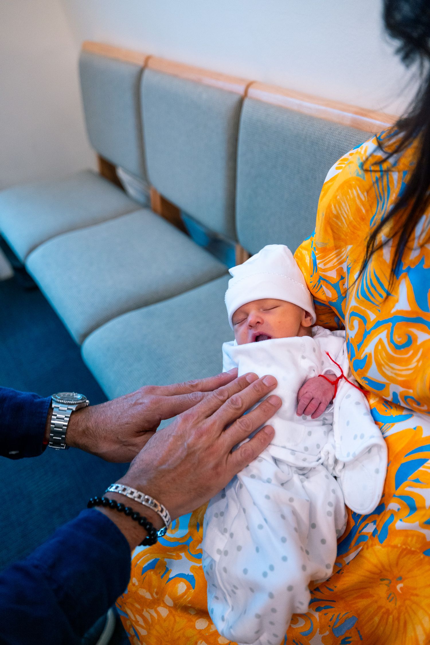 Close-up of hands tenderly holding a newborn baby wrapped in white clothing on blue chairs.