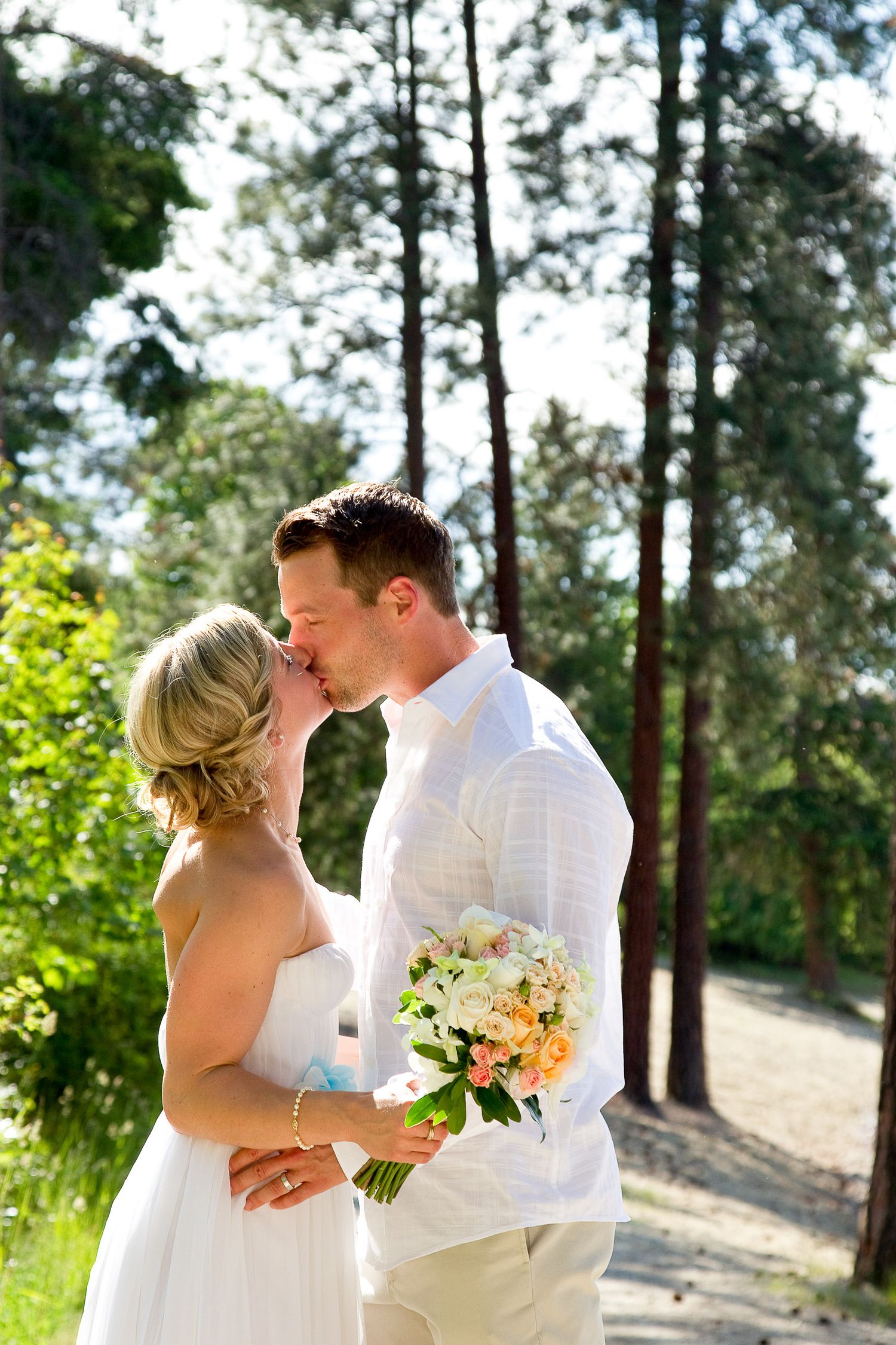 Wedding couple in white attire share romantic moments in a sunlit pine forest.