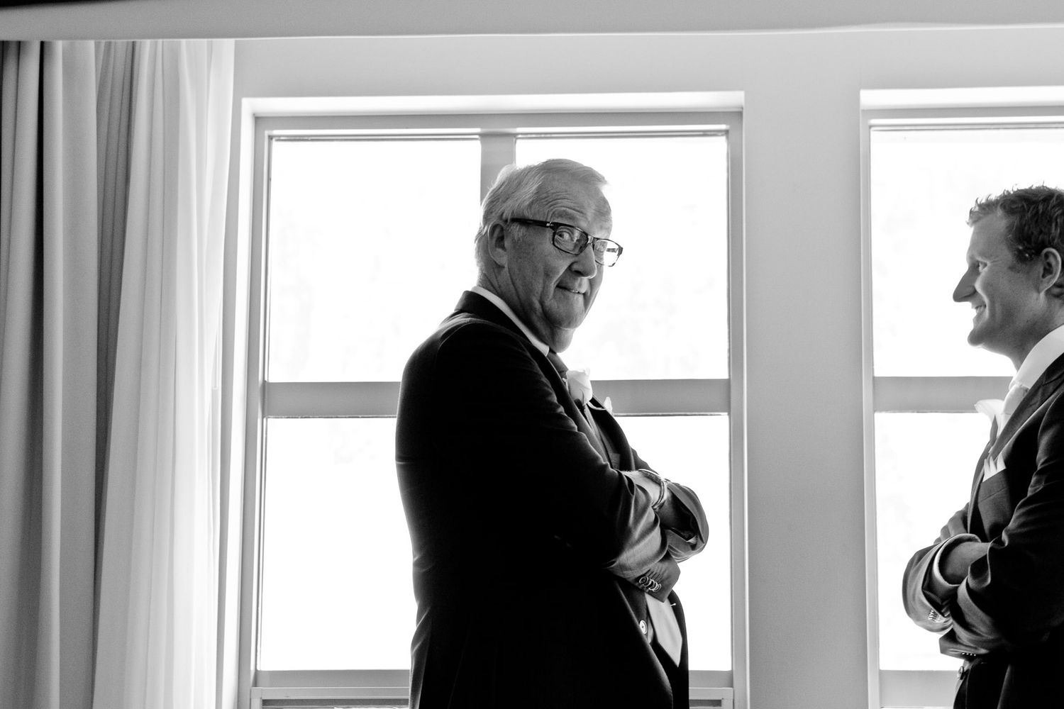 Two men in formal attire having a conversation by large windows in black and white.