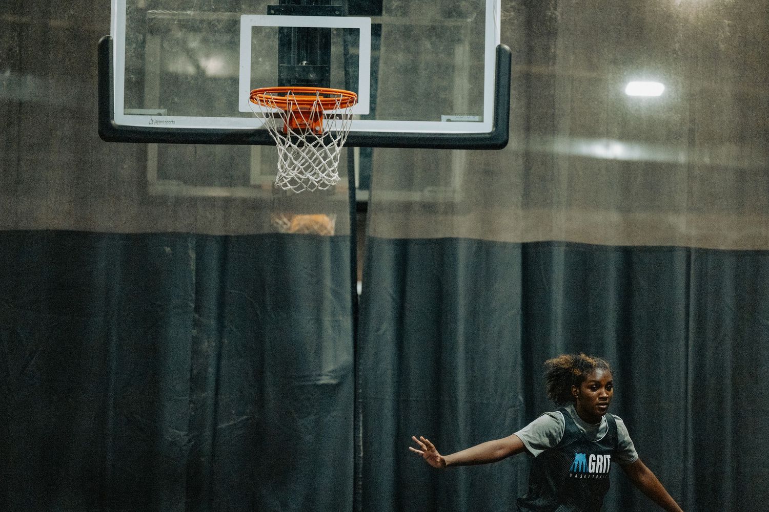 Player practices defensive stance beneath basketball hoop in indoor court.
