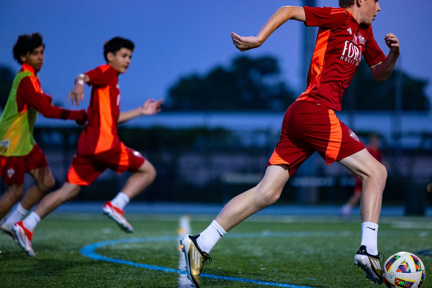 Athletes in red jerseys run and train on a soccer field during an evening practice session.