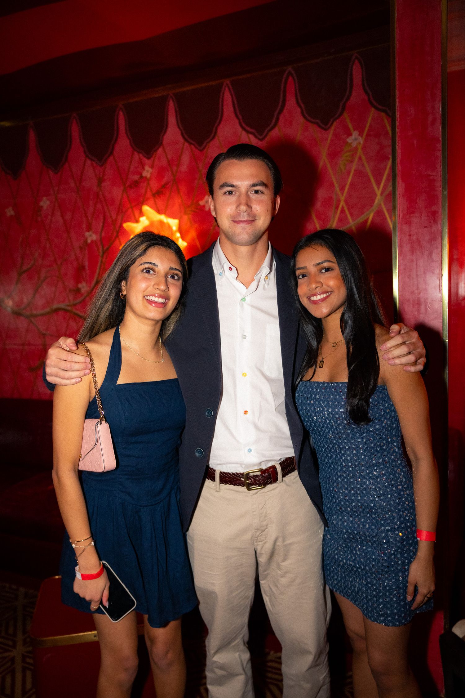 Three young people pose together against a red wall at an evening social event.