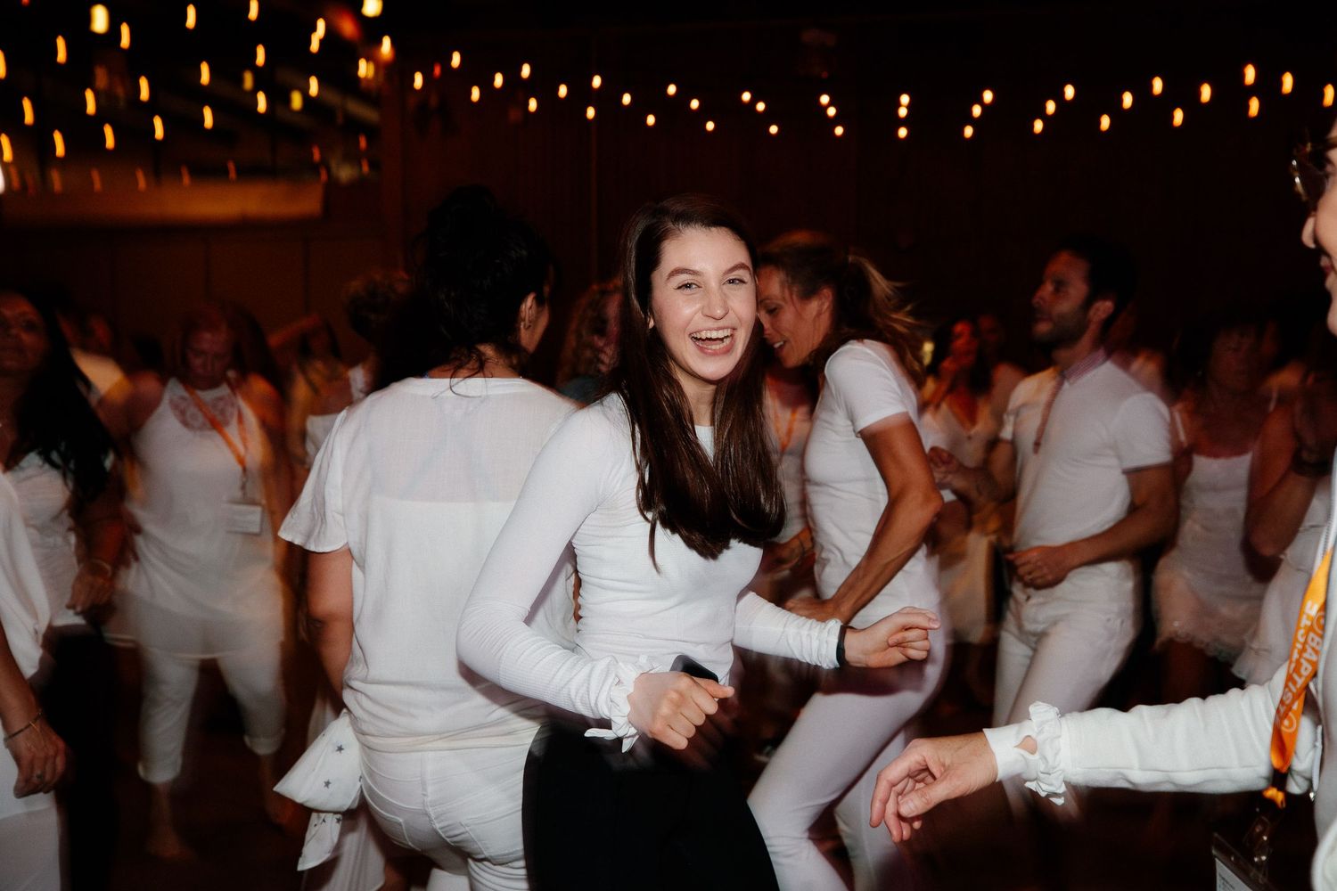 People in white outfits dance and celebrate under string lights at a nighttime party gathering.