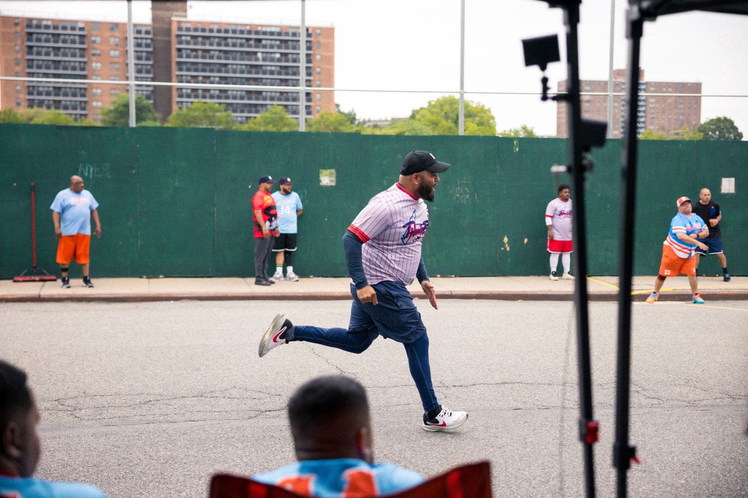 Baseball players warming up and practicing batting drills on an outdoor diamond with fences and buildings visible.