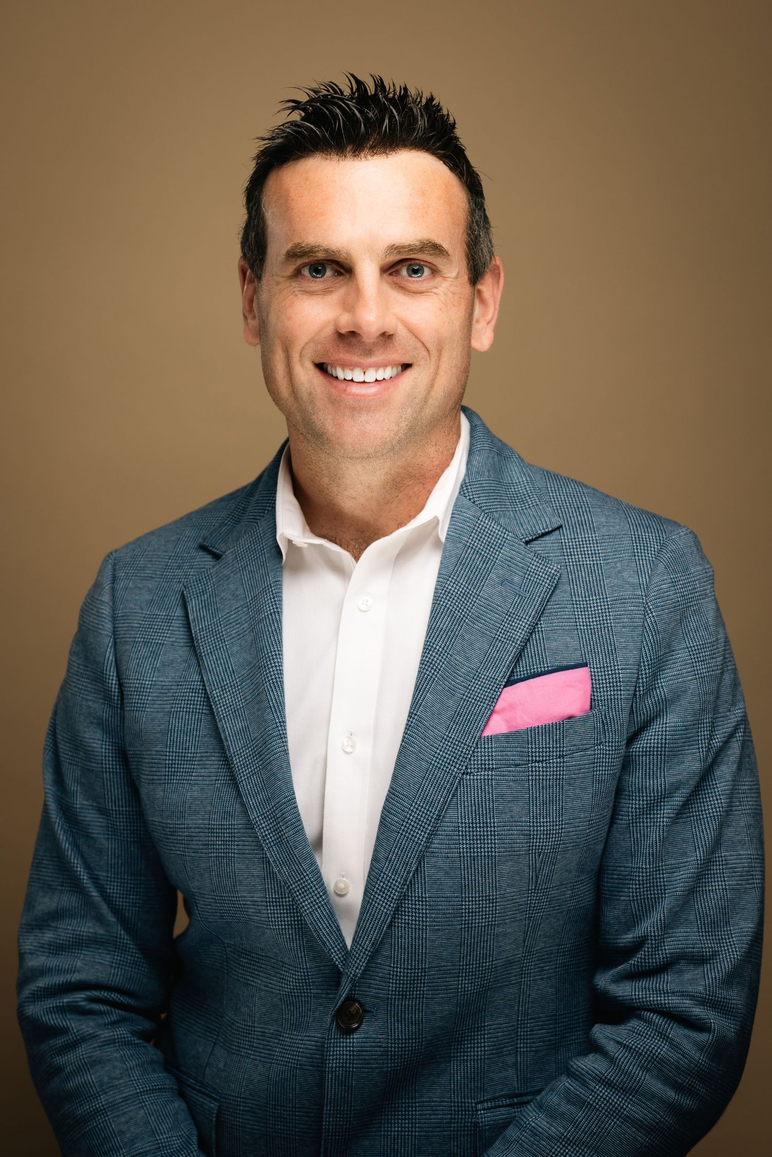 Smiling man in blue blazer with pink pocket square and white shirt posing against a warm beige background.