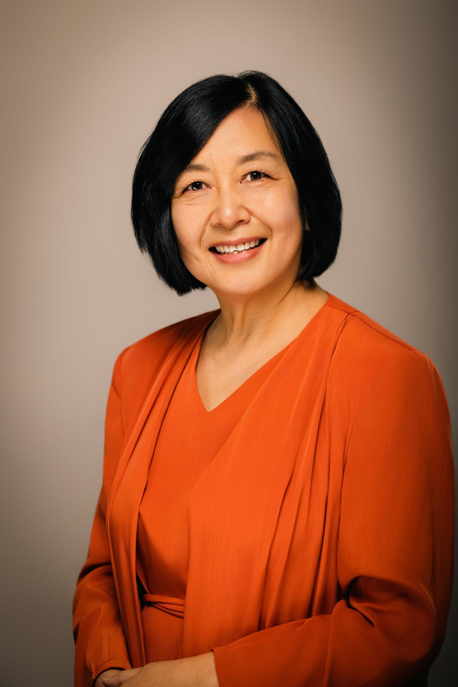 Smiling middle-aged Asian woman with black bob hair wearing an orange blouse, professional headshot portrait.