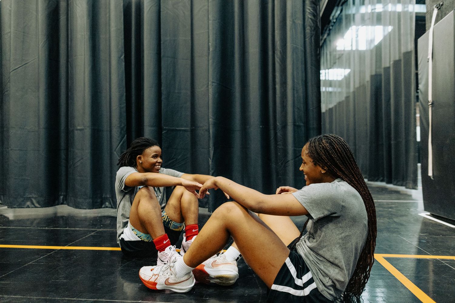 Two dancers resting together on the floor of a dance studio with black curtains and mirror walls.