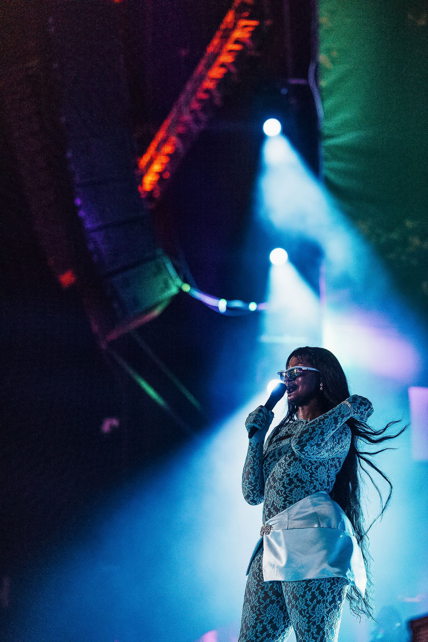 A performer in a patterned outfit stands on stage under dramatic blue spotlights during a concert performance.