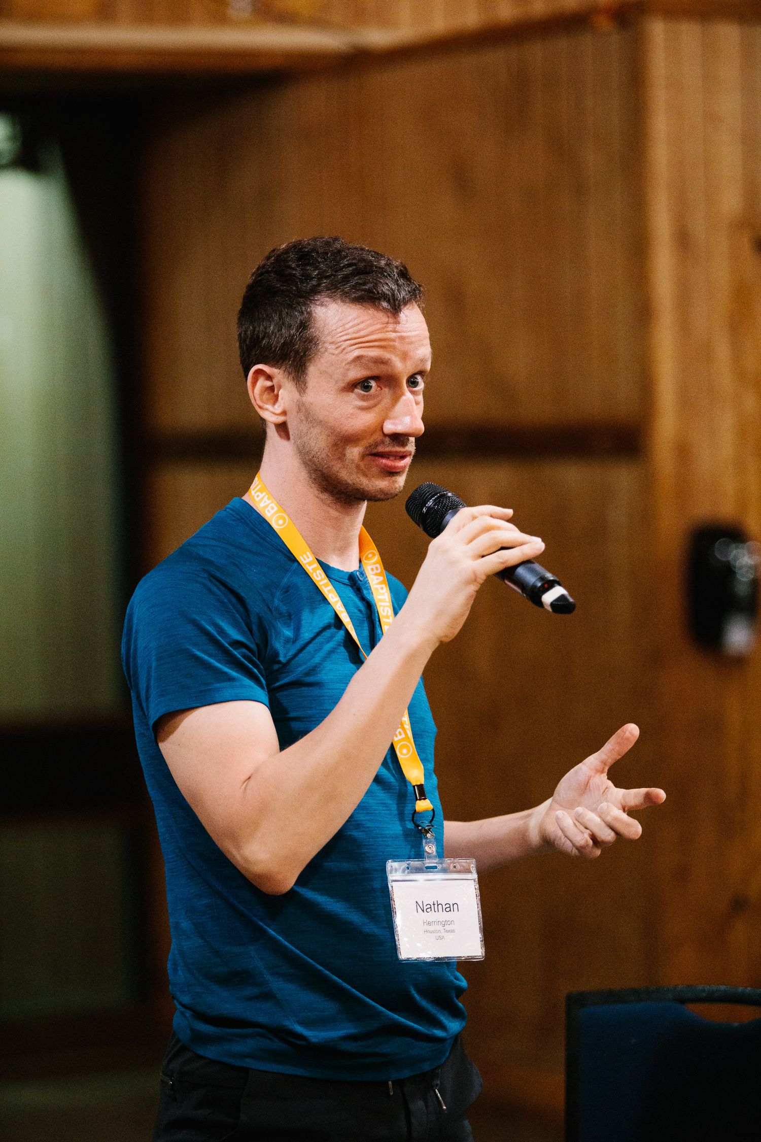 A speaker presents at a conference wearing a blue t-shirt and name badge in a wooden auditorium.