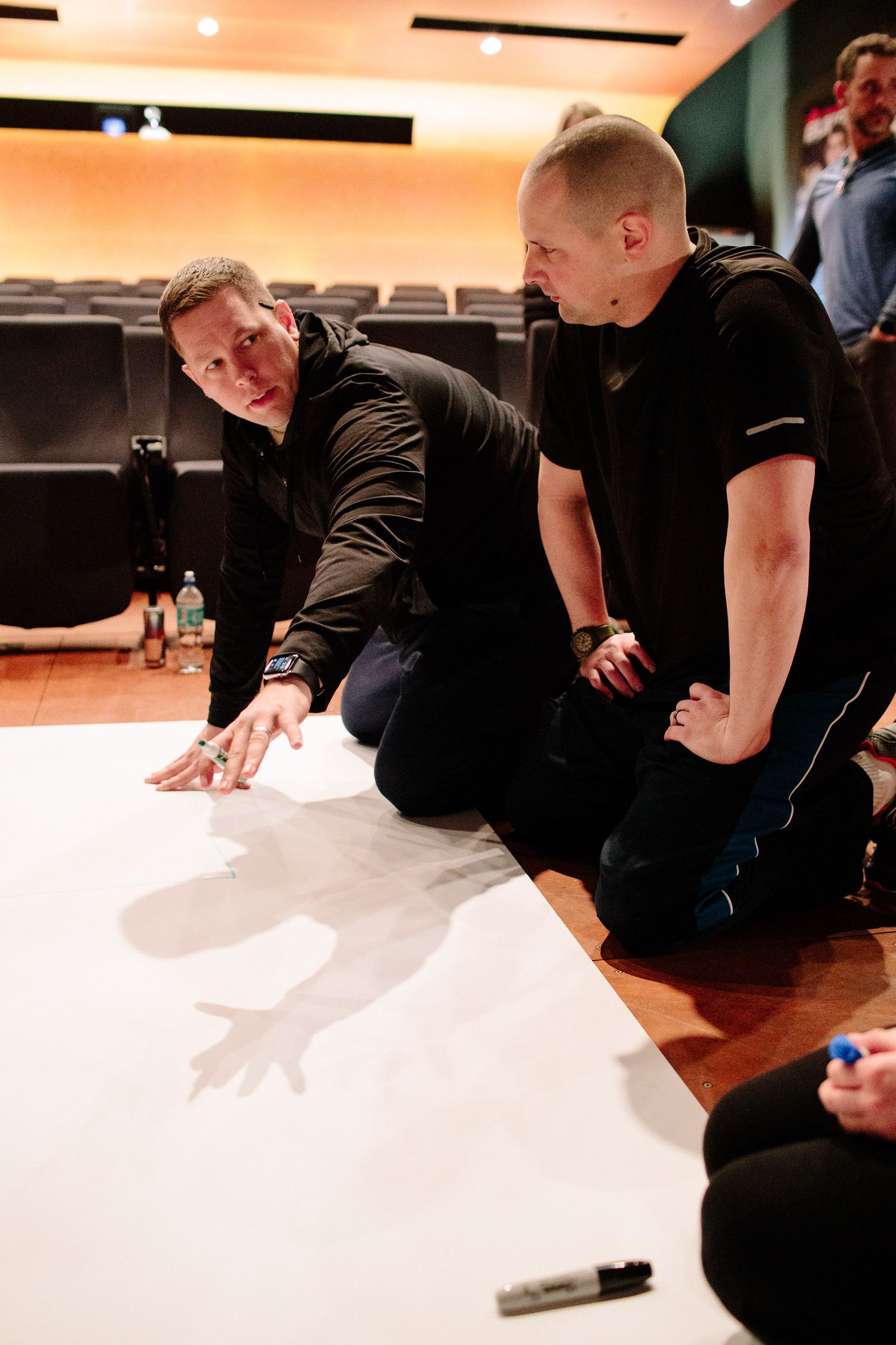 A group of performers rehearse movements while seated on a white floor.