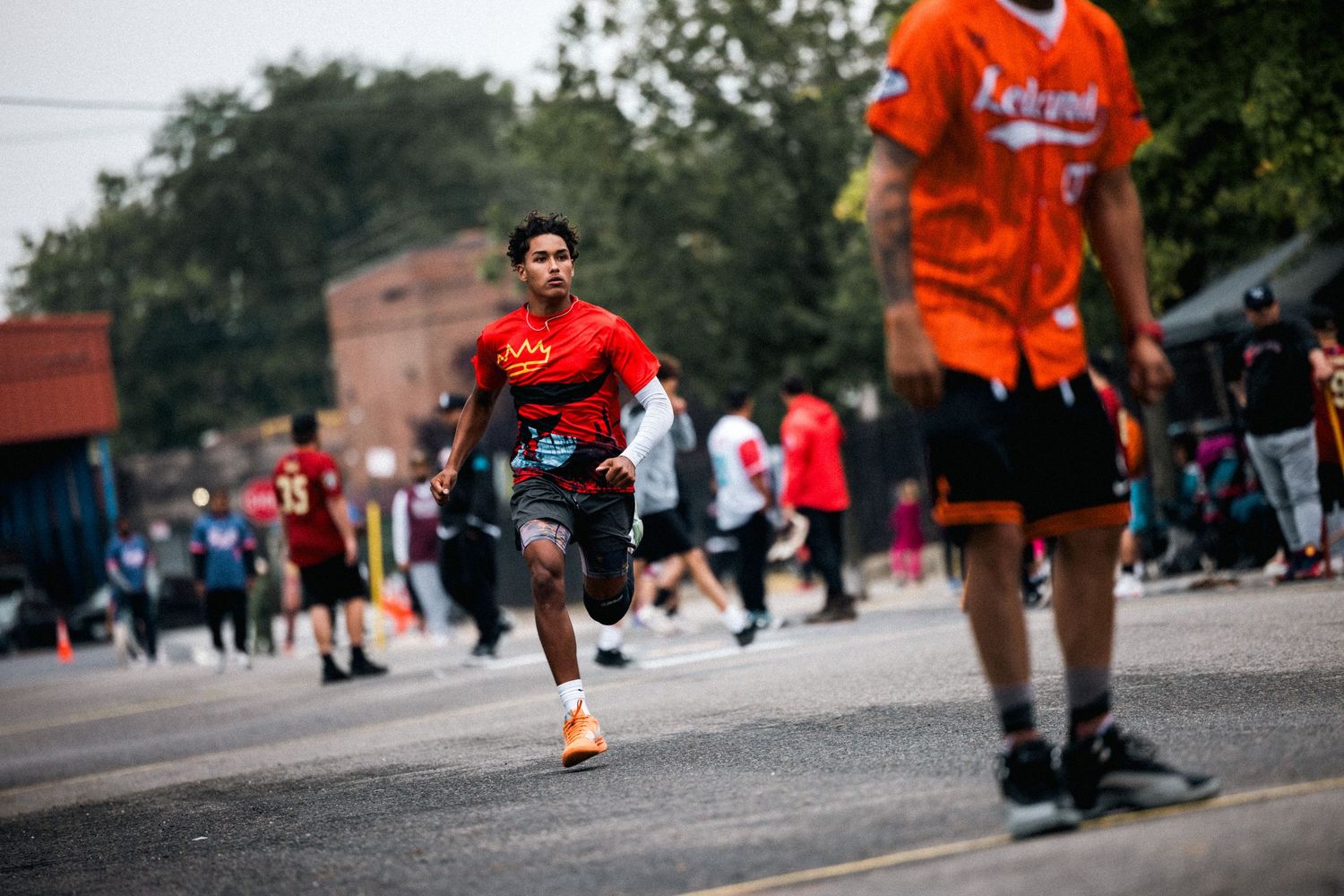 People in orange jerseys playing sports on an asphalt surface with spectators in the background.