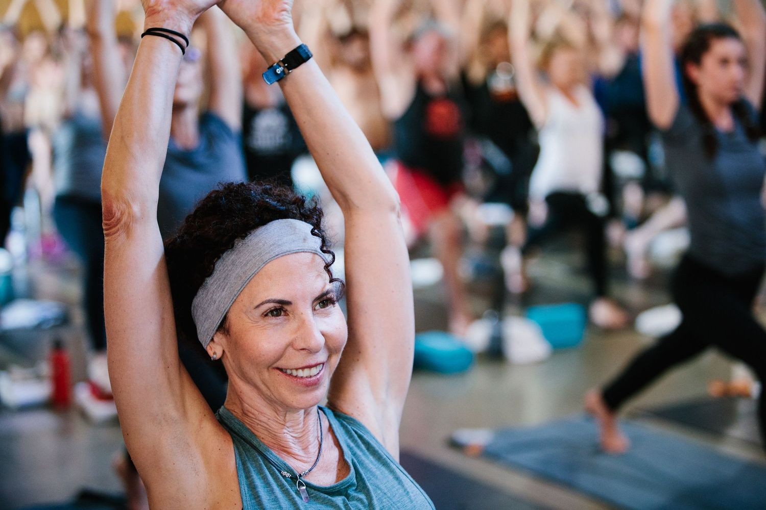 A group of people practice yoga together in a large studio, stretching their arms upward during class.