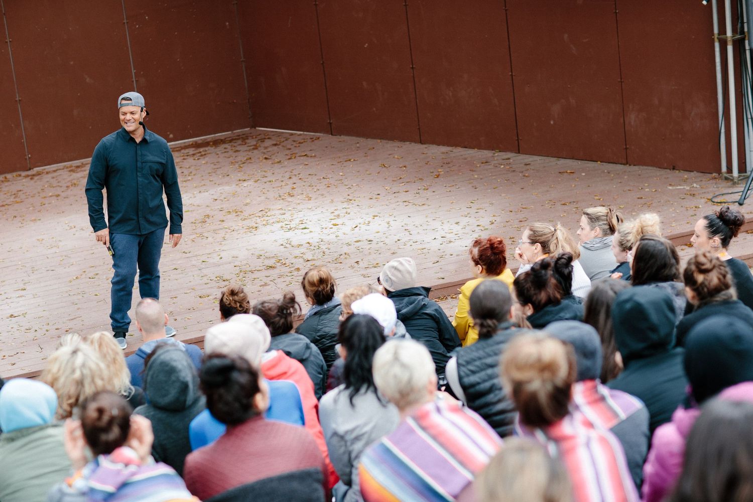 A speaker addresses a seated audience in an outdoor amphitheater with rustic metal walls.