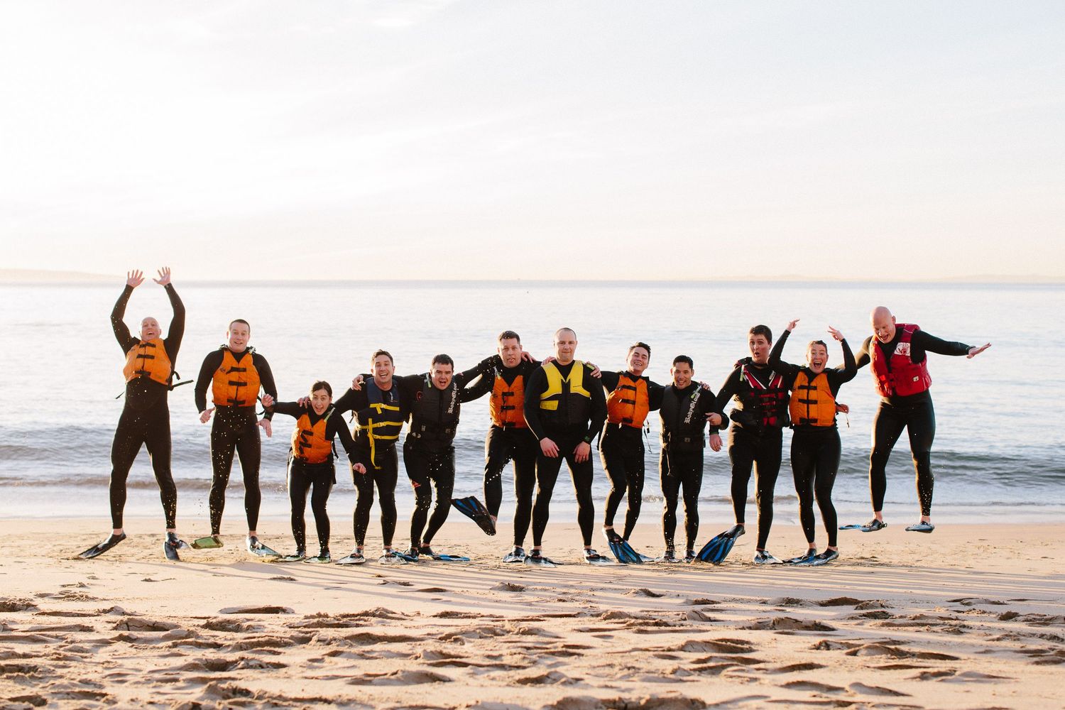 Group in wetsuits jumps joyfully on beach at sunset with ocean in background.