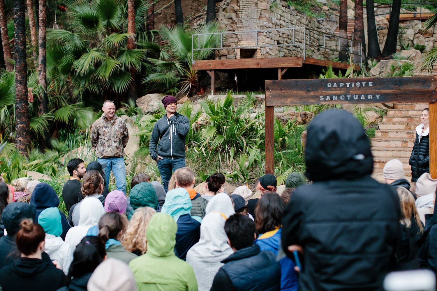 Outdoor gathering with attendees seated in rows among pine trees.