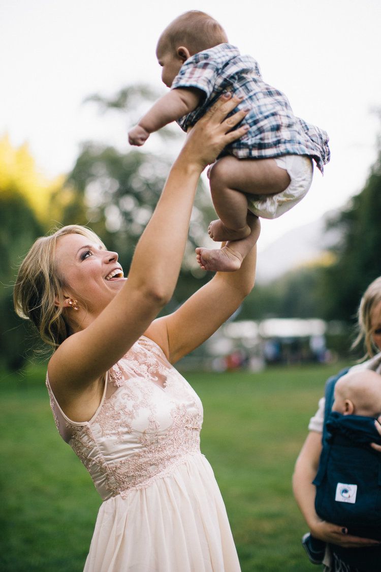 Woman in floral dress joyfully holds up baby wearing plaid in grassy field.
