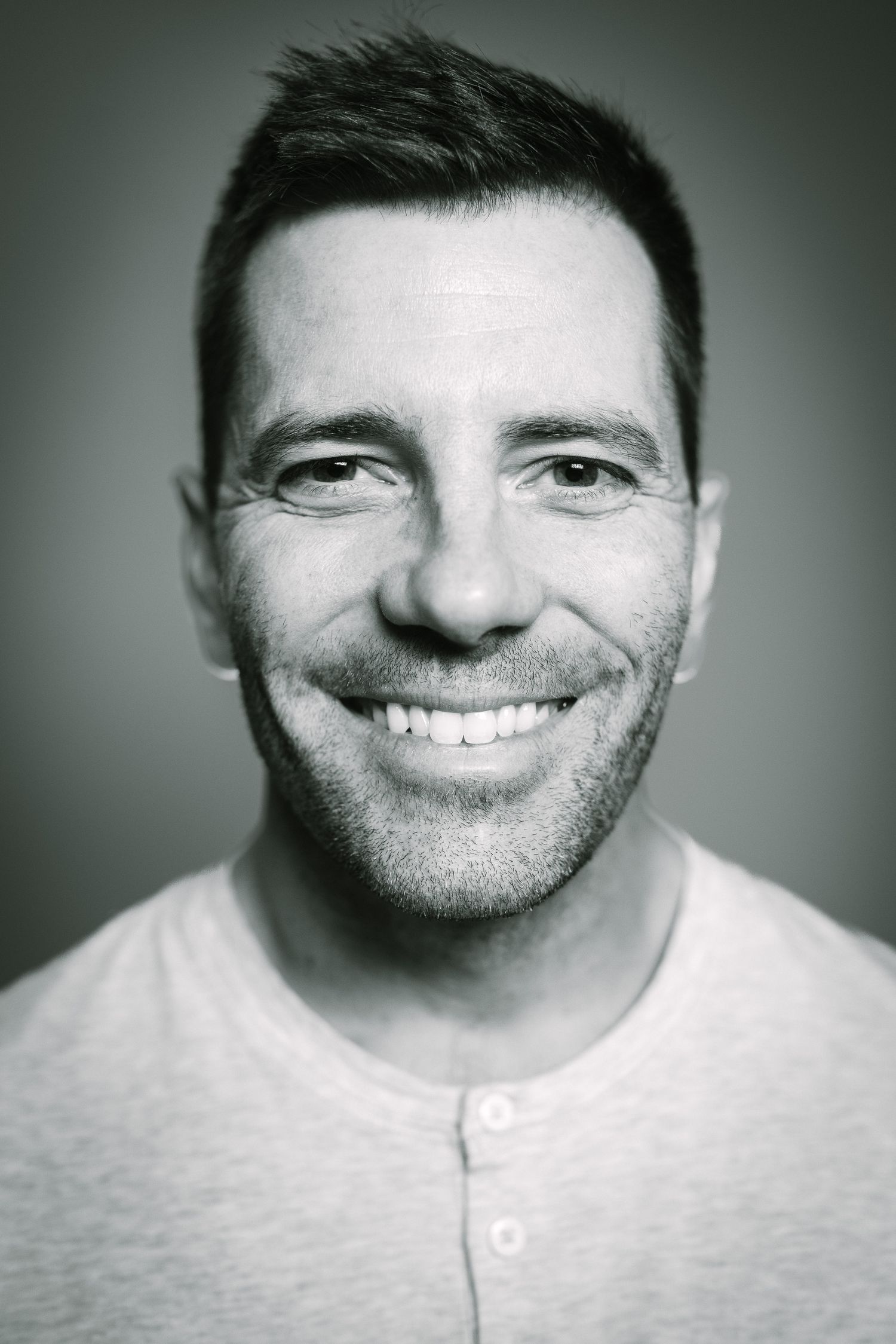Black and white portrait of a smiling young man with stubble wearing a henley shirt.