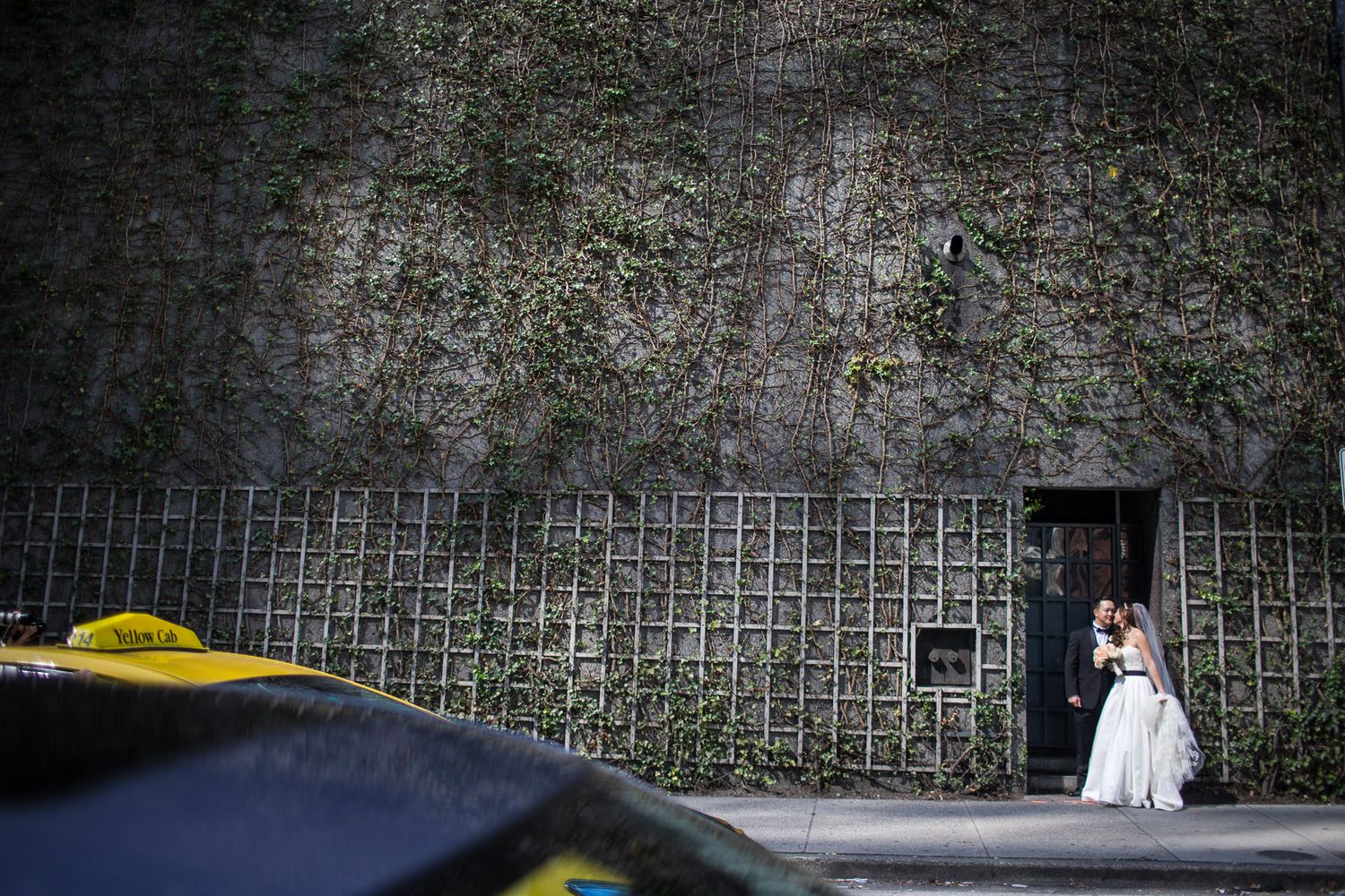 A bride in white poses against an industrial wall as a yellow taxi passes by.