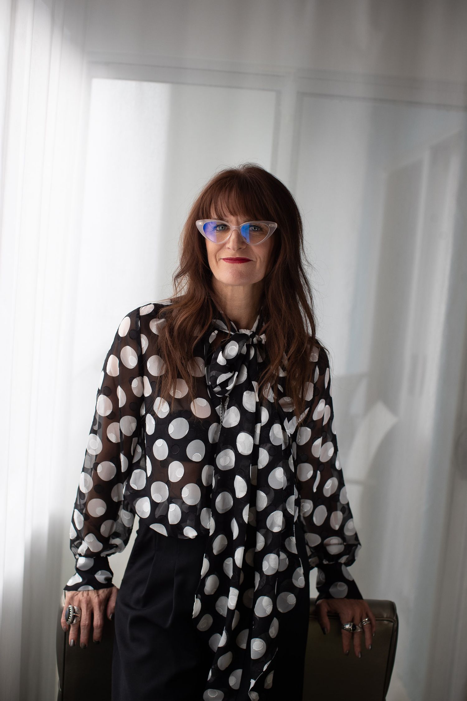 A person wearing polka dot blouse and blue glasses poses against white curtained background.