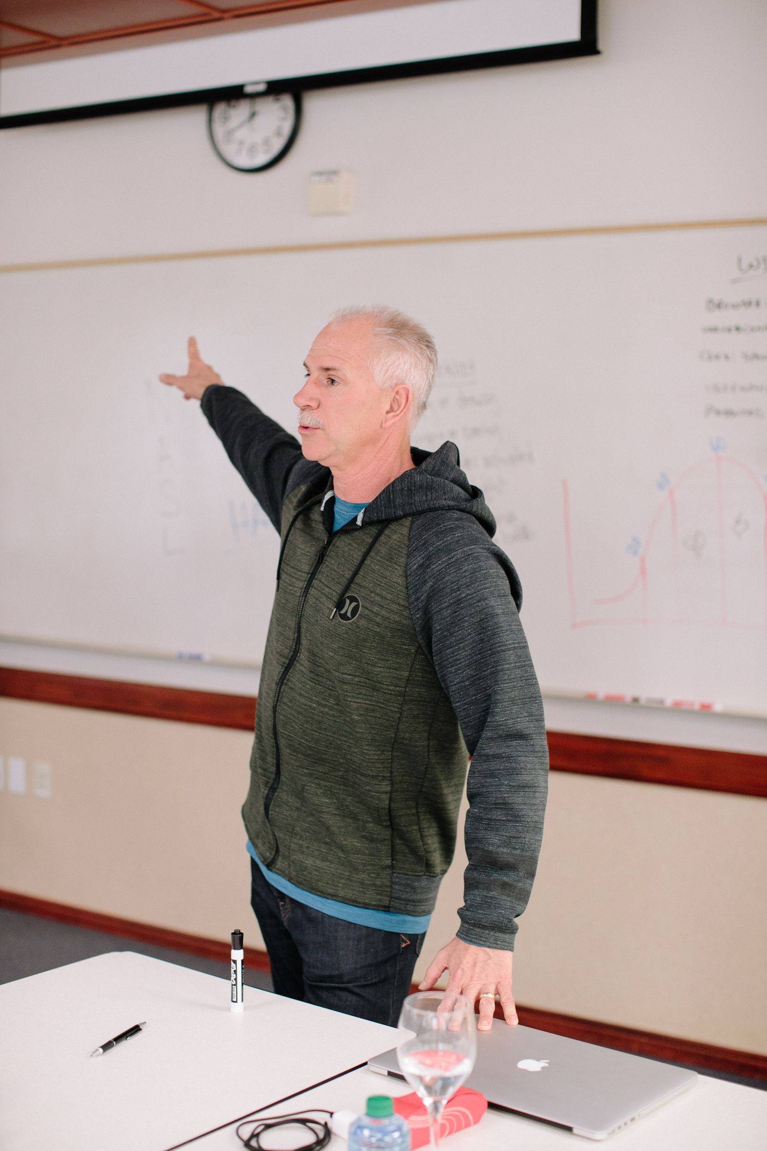 Someone in a grey hoodie gestures while teaching in front of a whiteboard with a water glass nearby.