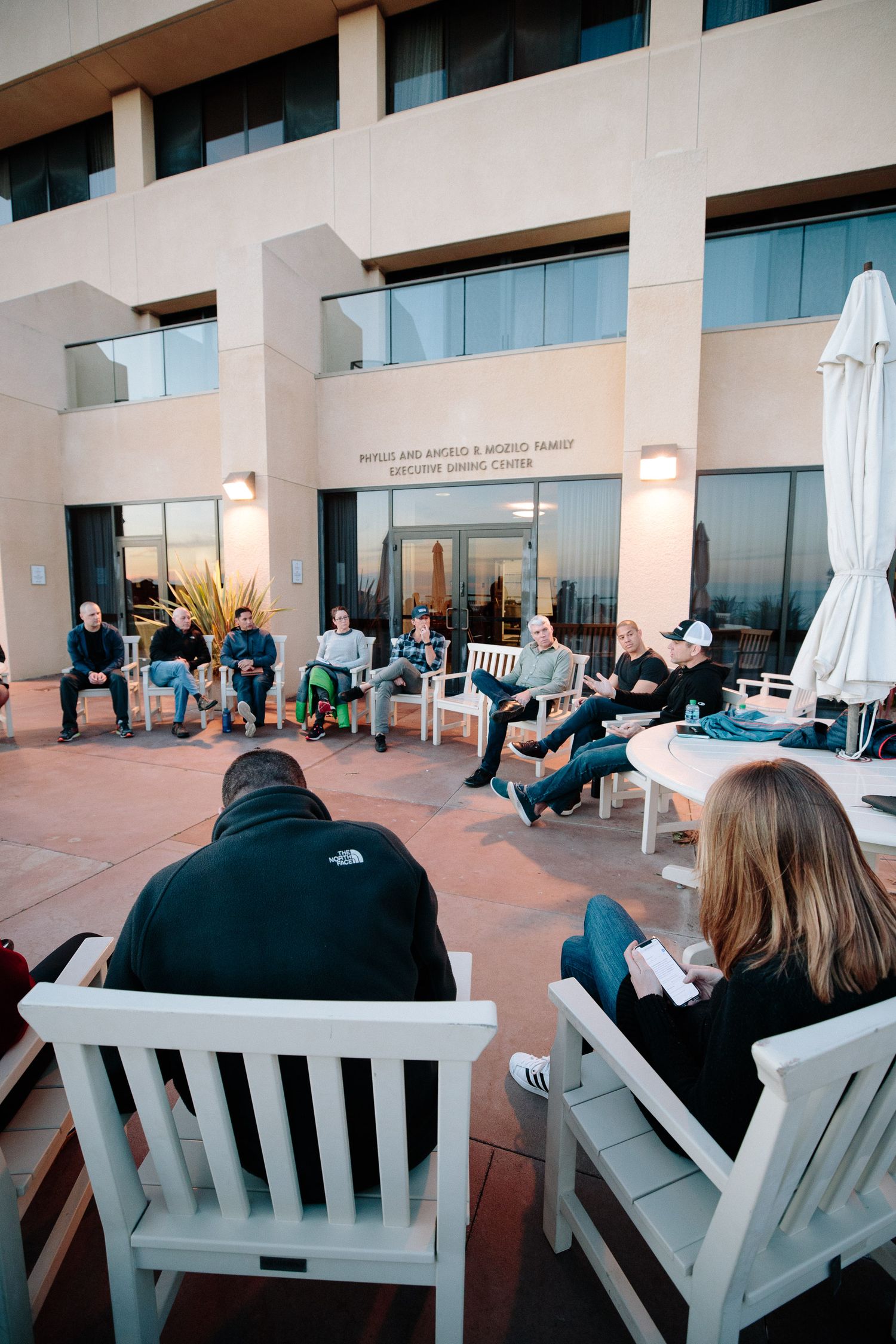 Attendees sit in white Adirondack chairs on an outdoor patio during a gathering.