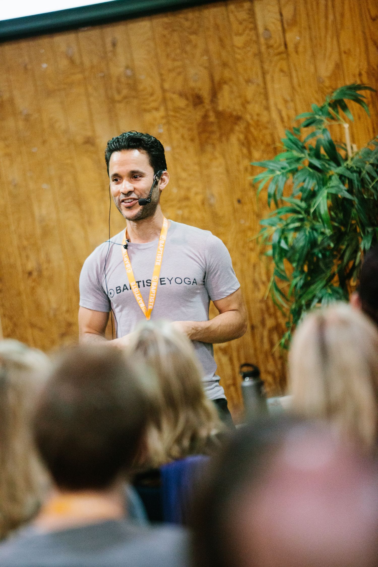 Speaker presenting to an attentive audience in front of a wood-paneled wall with green plants in the background.