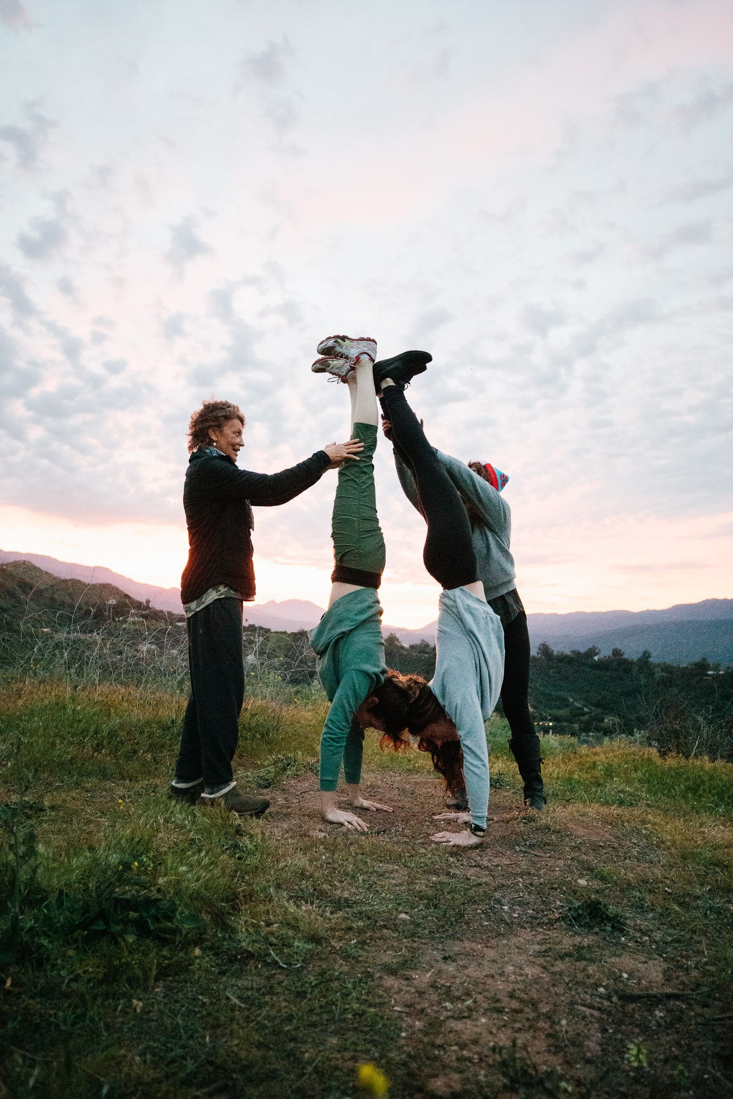 Three people performing acrobatic moves against a sunset mountain backdrop.