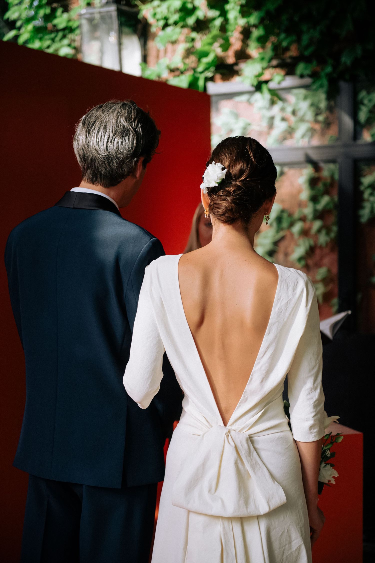 Back view of a couple in formal attire as the bride's white dress with bow detail contrasts against a dark background.