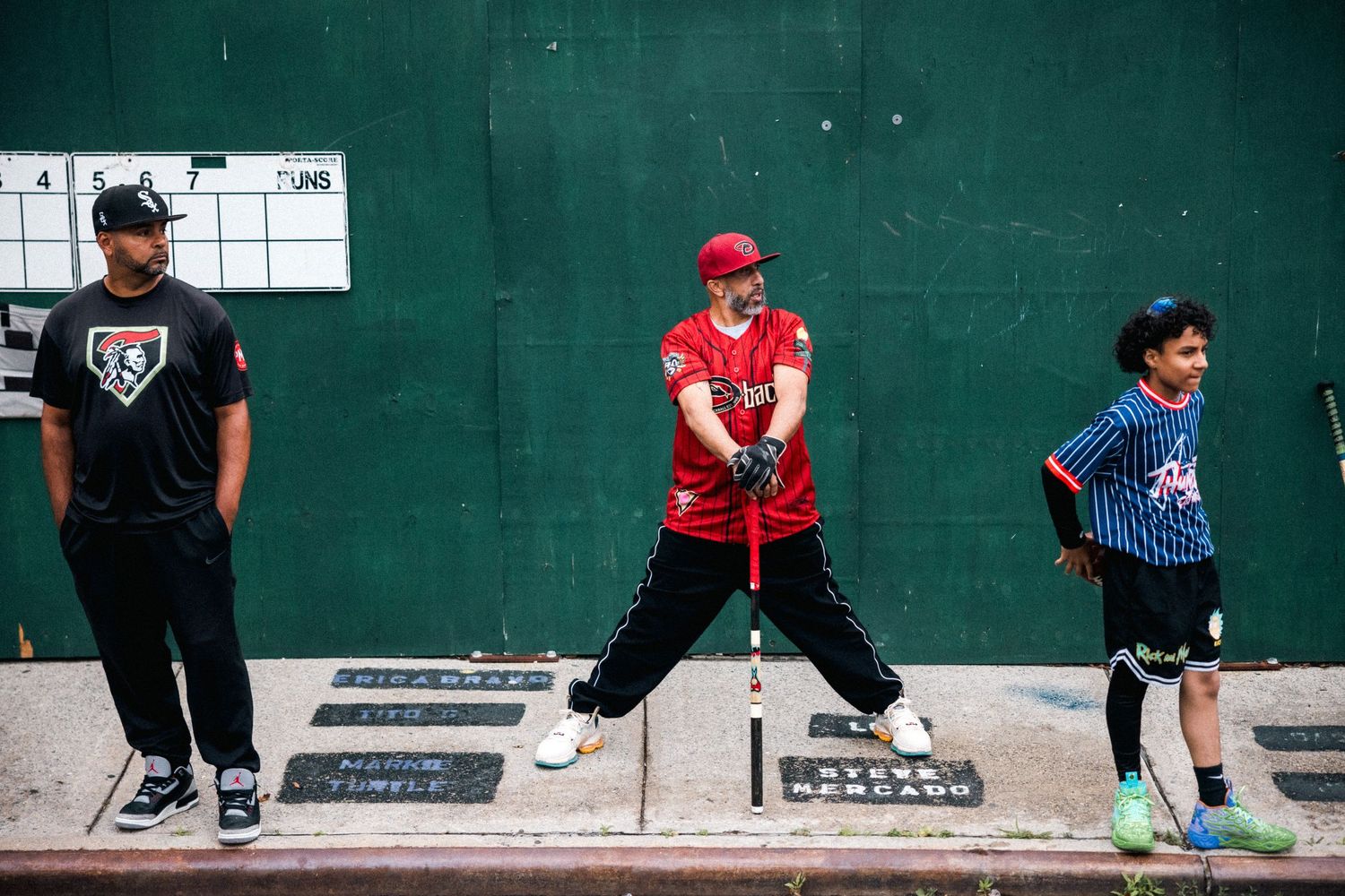Three people in casual athletic wear stand in a batting lineup along a concrete wall at a batting cage facility.