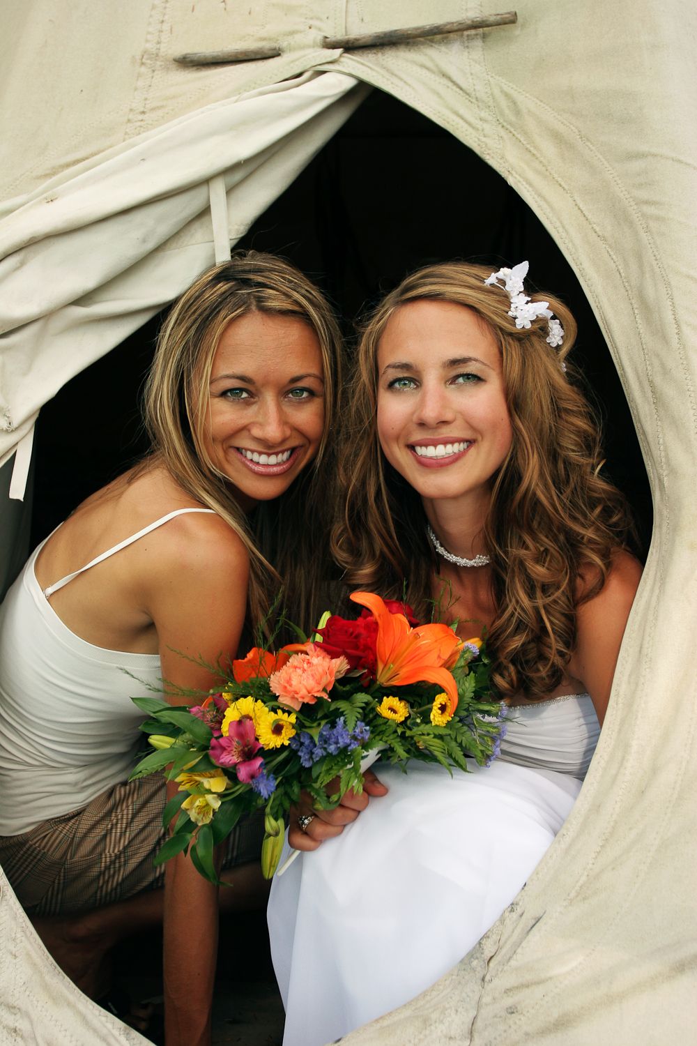 Two people in white dresses pose with colorful bouquets under a fabric canopy.