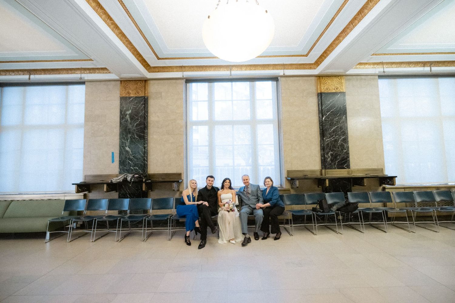 Small wedding party posing together in an elegant courthouse hall with marble columns and blue chairs.