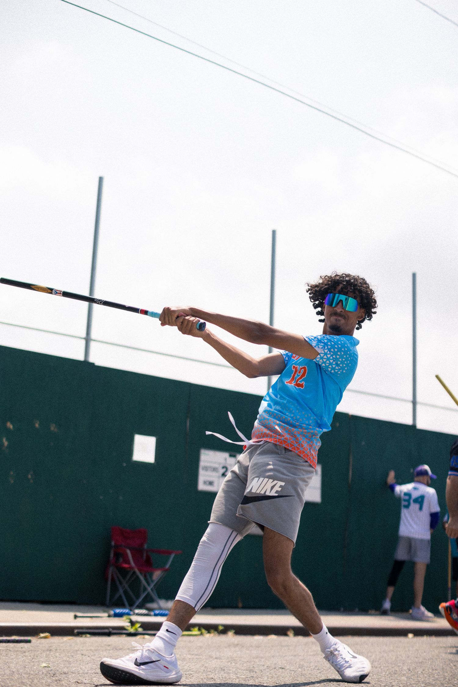 Baseball player in blue jersey and grey shorts swings bat during outdoor batting practice against green wall backdrop.