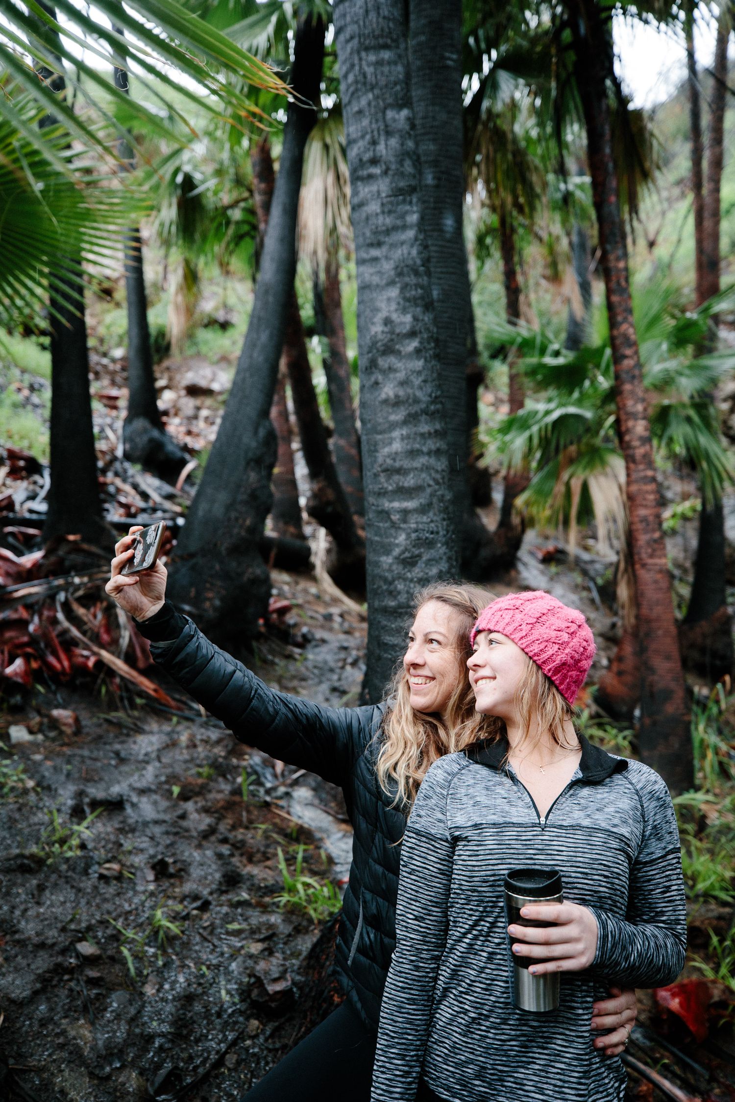 Two people share a playful moment in a palm tree forest, wearing casual outerwear and a red knit hat.