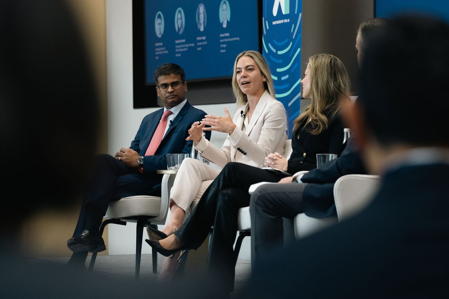 Business professionals participate in a panel discussion at a conference with blue digital displays in background.