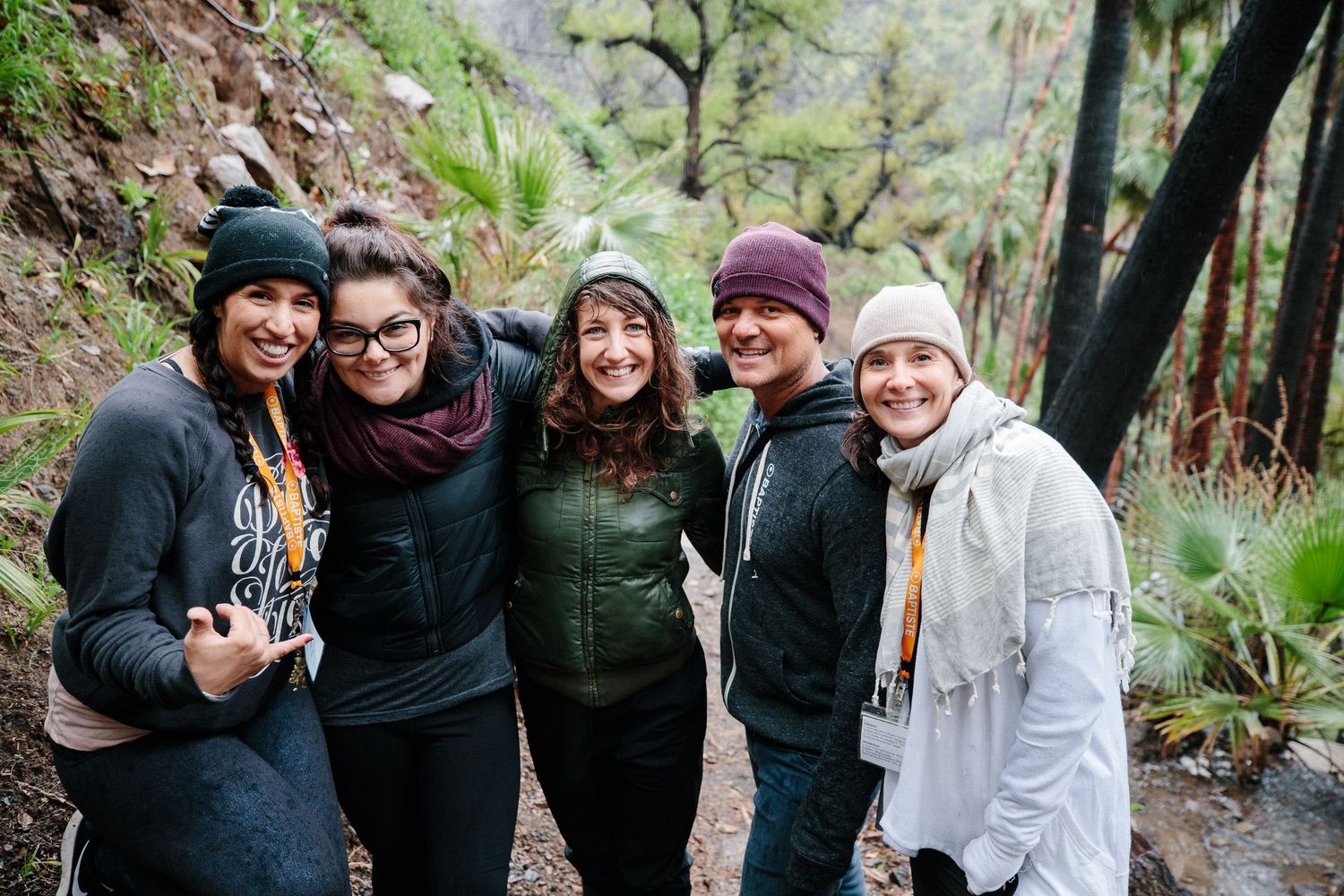A group of friends wearing winter clothing and beanies smile together on a hiking trail surrounded by lush greenery.
