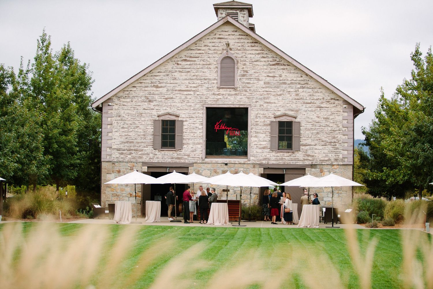 White stone barn building with outdoor seating area framed by green grass and trees.