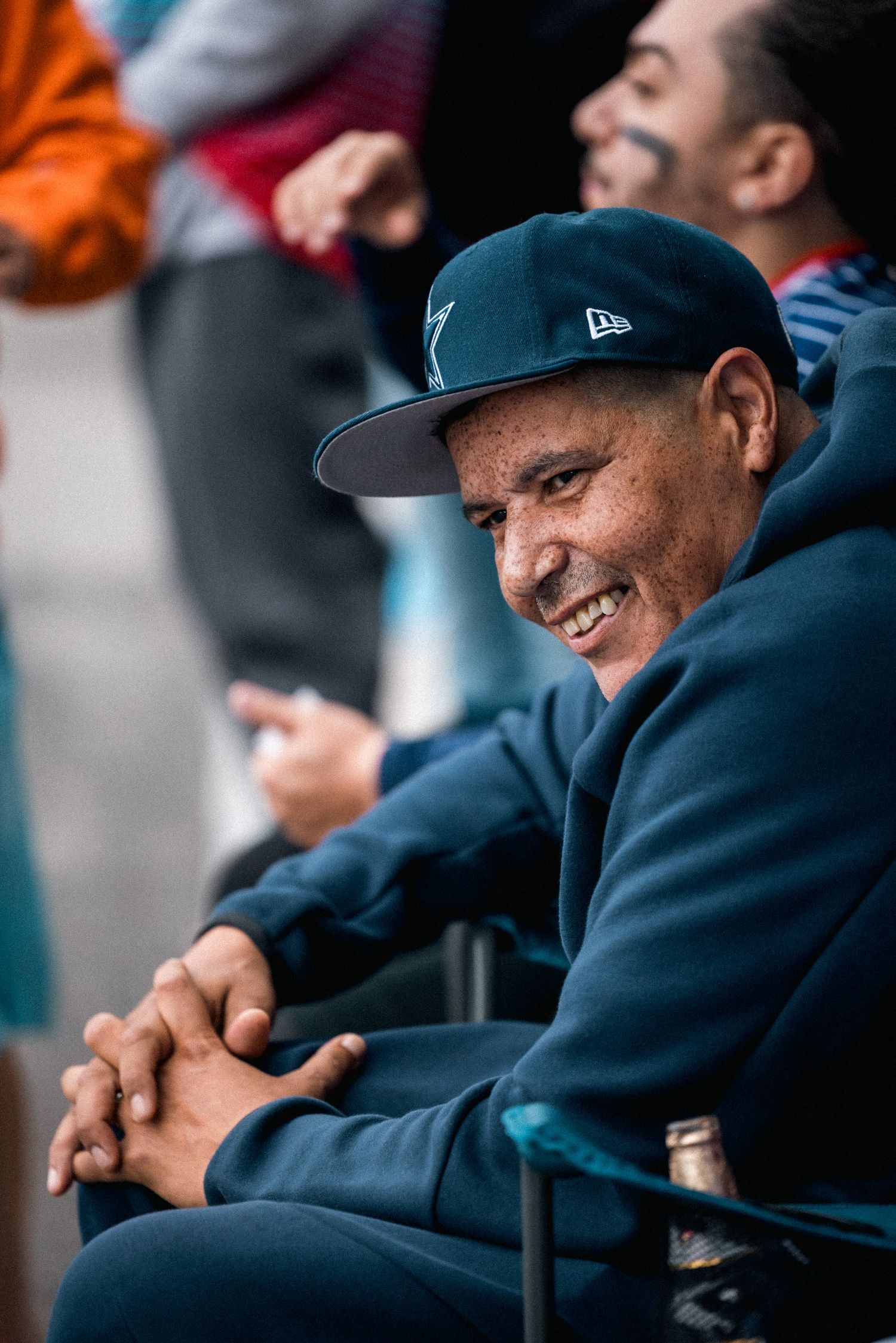 A person in a New Era baseball cap and navy jacket smiles while seated at an event.