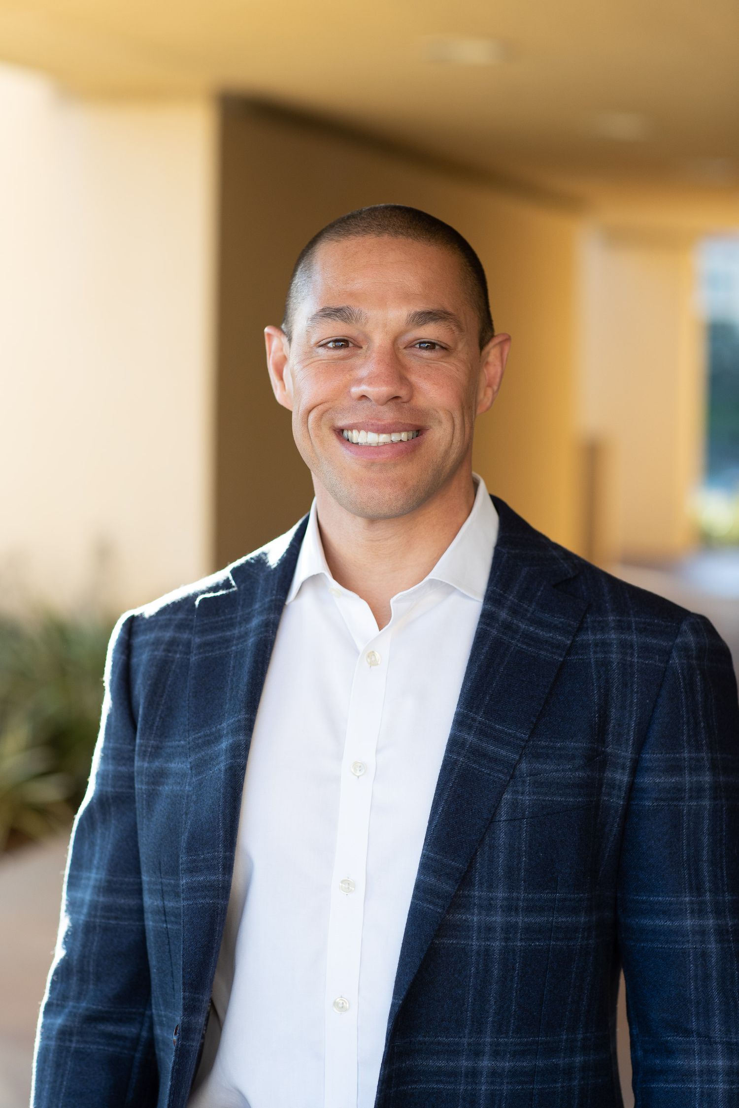 A professional wearing a navy plaid blazer and white shirt smiles in a hallway setting.