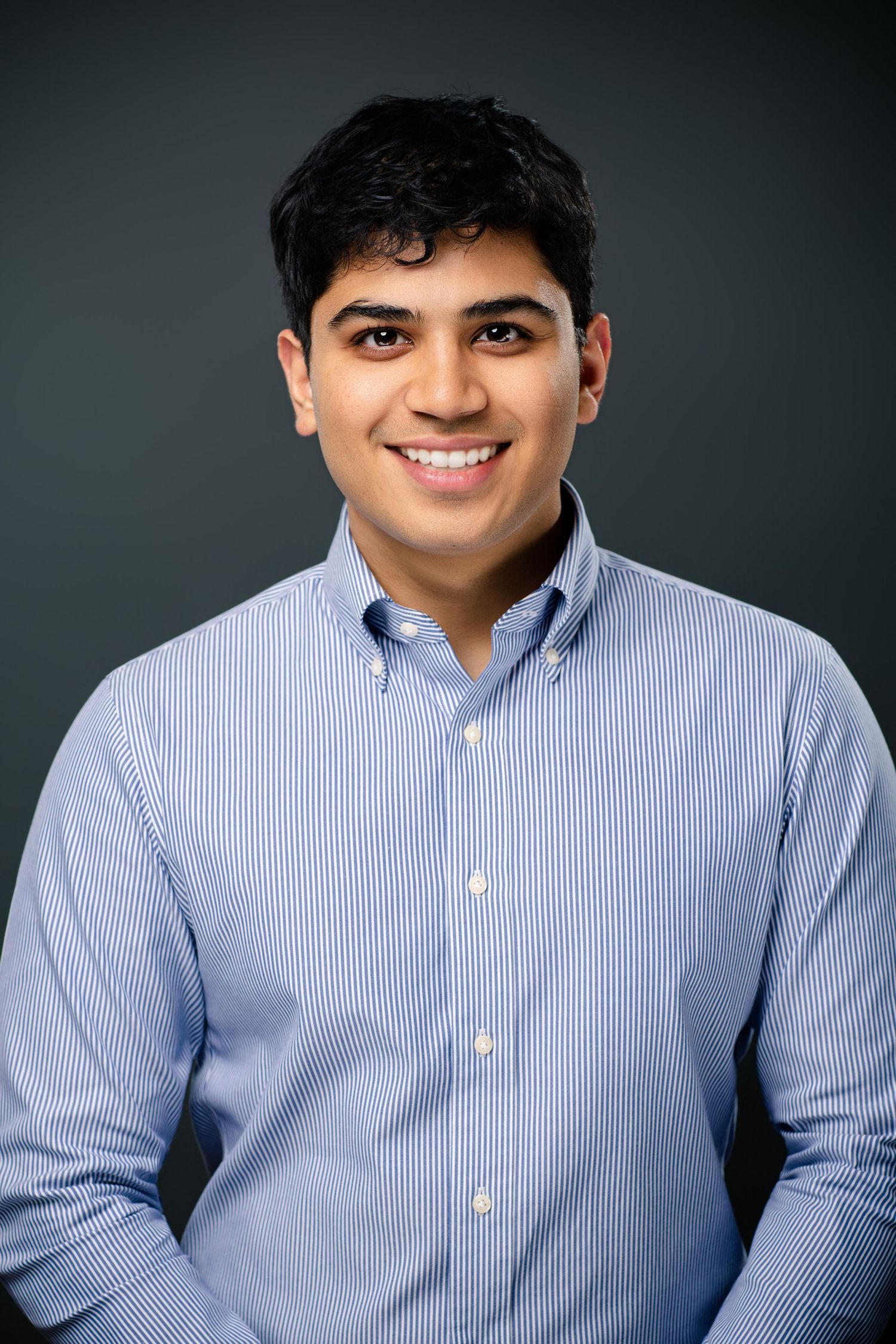 Young man in a light blue striped button-up shirt smiling confidently against a dark gray background.